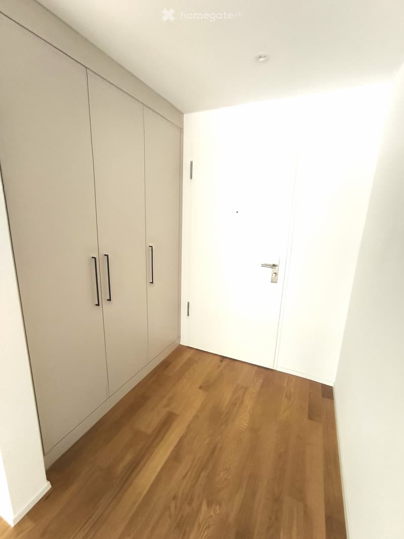 A clean, minimalist hallway with light wooden flooring, white walls, built-in storage units with black handles, and a closed white door at the end.