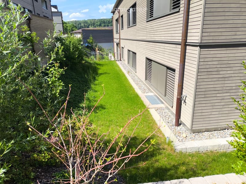 A narrow, well-maintained garden area with a small patch of green grass, surrounded by gravel pathways and wooden fences. The garden is bordered by shrubs and trees, with a modern house visible on the right side.