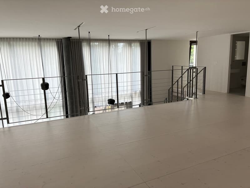 Empty room with wooden floor, large windows with curtains, metal railing, and a staircase leading to another level.