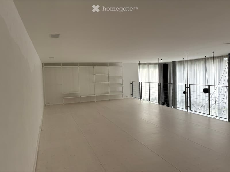 Empty room with light-colored tiled floor, white walls, and a large window with sheer curtains. A metal shelving unit is mounted on the wall, and a black metal railing is visible near the window.