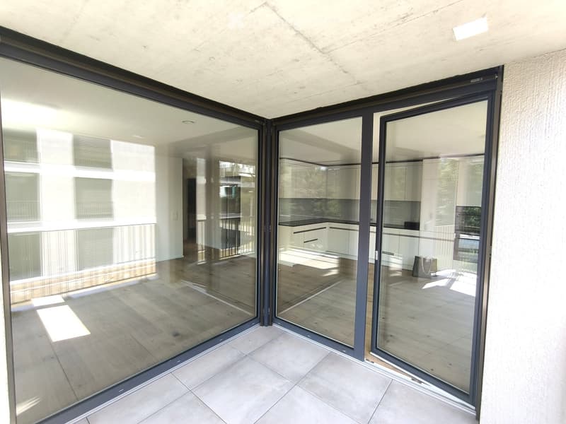 Modern balcony with large glass sliding doors, tiled floor, and a view into a contemporary kitchen with white cabinets and dark countertops.