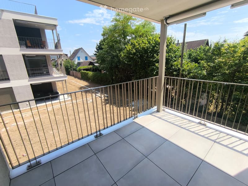 Balcony with tiled floor and metal railing, overlooking a landscaped area with trees and neighboring houses.