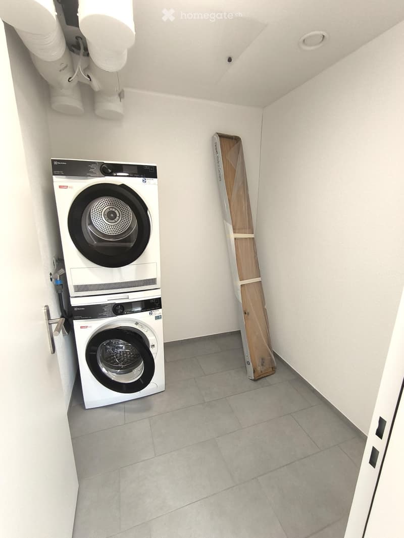 A small, clean room with a white washing machine and dryer unit against the wall, tiled floor, and a wooden packing box on the floor.