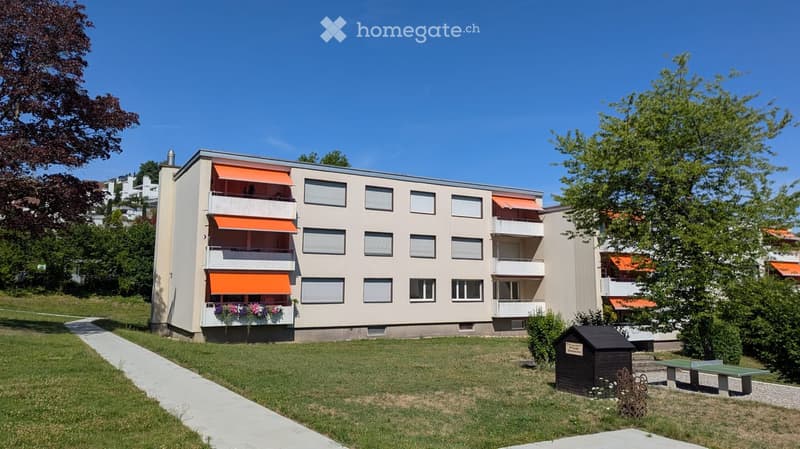 Modern multi-story residential building with balconies, orange awnings, and flower boxes. The surrounding area includes a lawn, pathway, and outdoor seating area with a small shed and picnic tables.