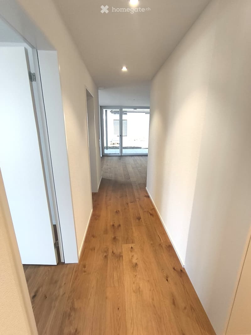 Empty hallway with wooden flooring, white walls, and recessed lighting. Features a glass door at the end leading to another room.