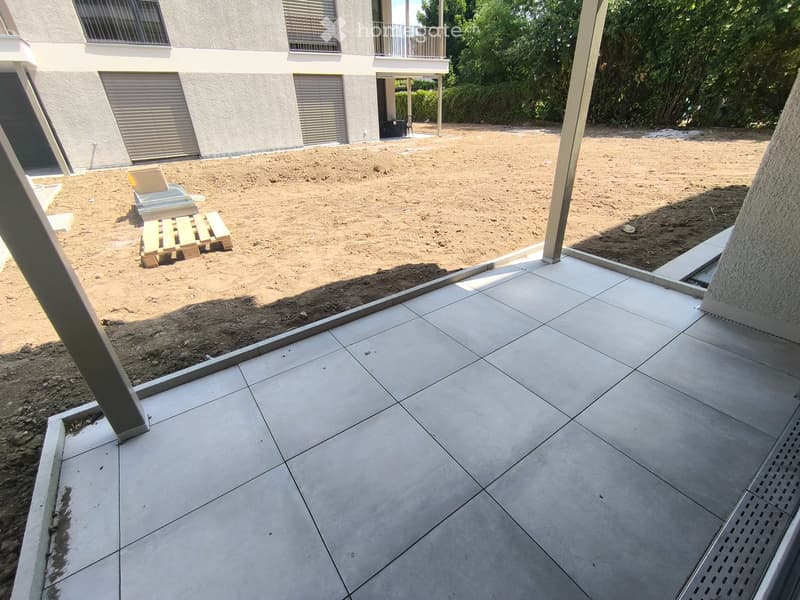 Outdoor area with sandy ground, wooden pallets, and a tiled patio adjacent to a building with balconies and windows.