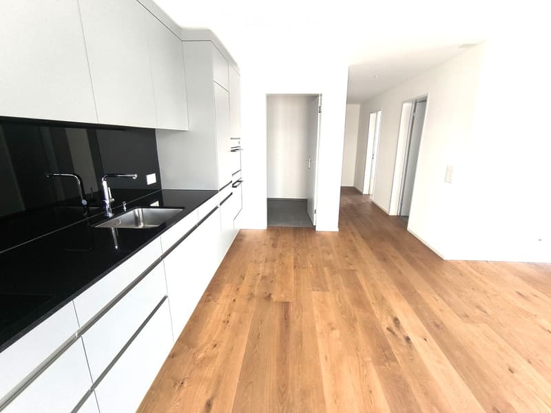 Empty room with light wooden flooring, white walls, and a modern kitchen area featuring white cabinets, a black countertop, and a stainless steel sink.