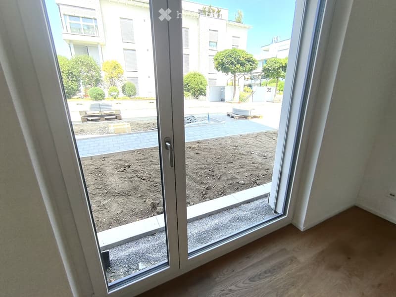 Empty room with wooden flooring and a large glass sliding door leading to an outdoor area with dirt ground and a paved walkway.