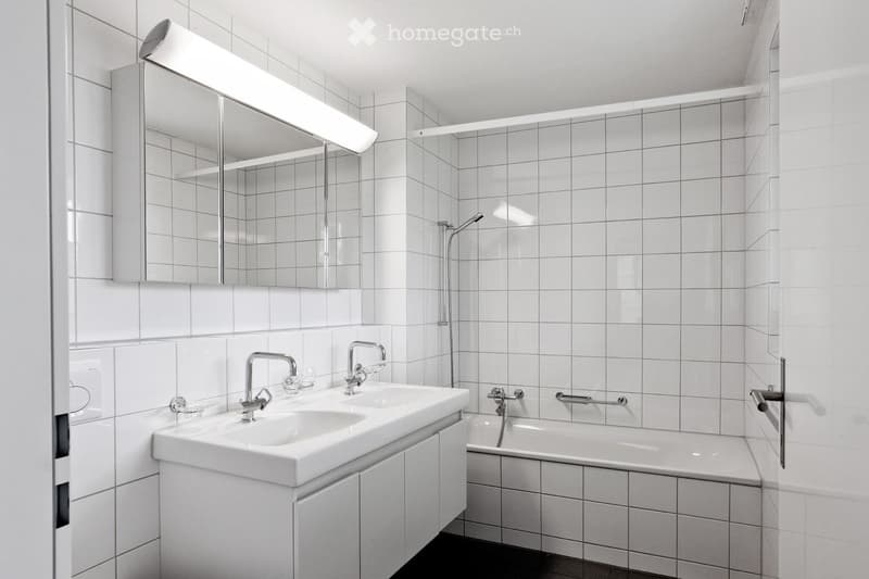 Tiled walls and floors, white fixtures, shower, bathtub, double sink with cabinets, mirror with integrated light