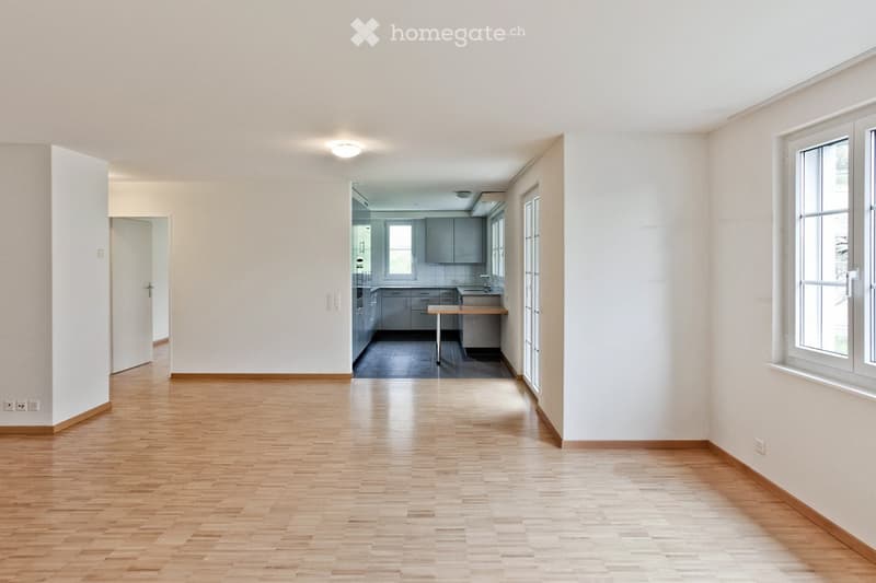 Empty room with wooden floors, white walls, window, kitchen is visible through the corridor