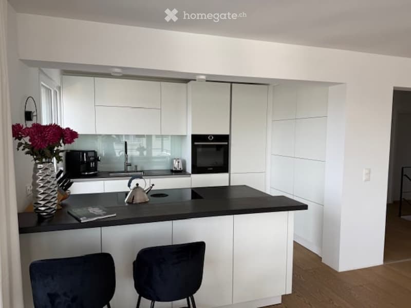 Modern kitchen with white cabinets, black countertop, coffee maker, oven, faucet, vase with flowers, and two chairs.