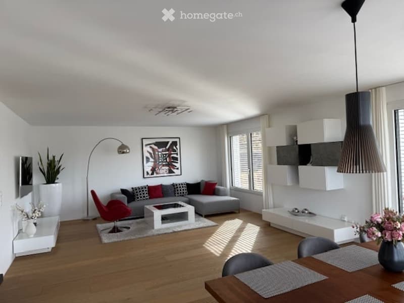 Modern living room with grey sectional sofa, red accent chair, white coffee table, wooden floor, large windows, and decorative elements including a floor lamp and potted plants.