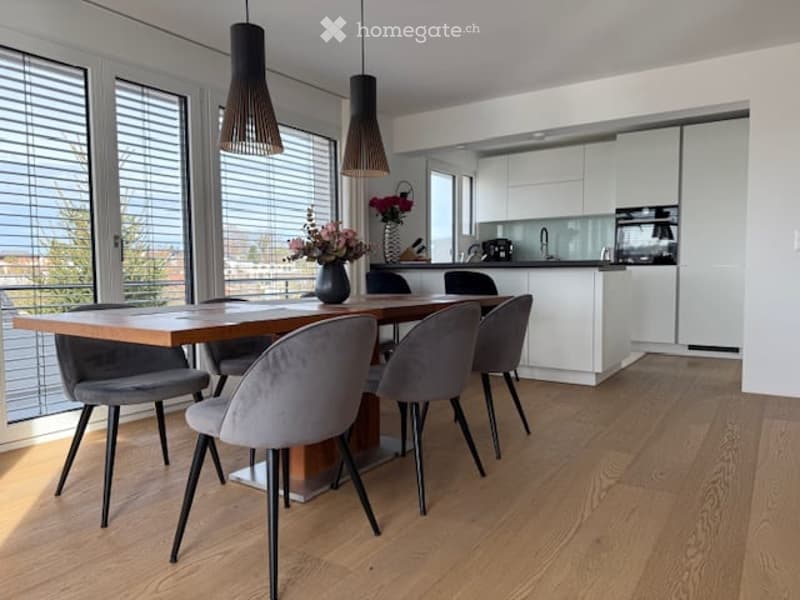 Modern dining area with a wooden dining table and grey chairs, a vase with flowers on the table, and a sleek white kitchen in the background.