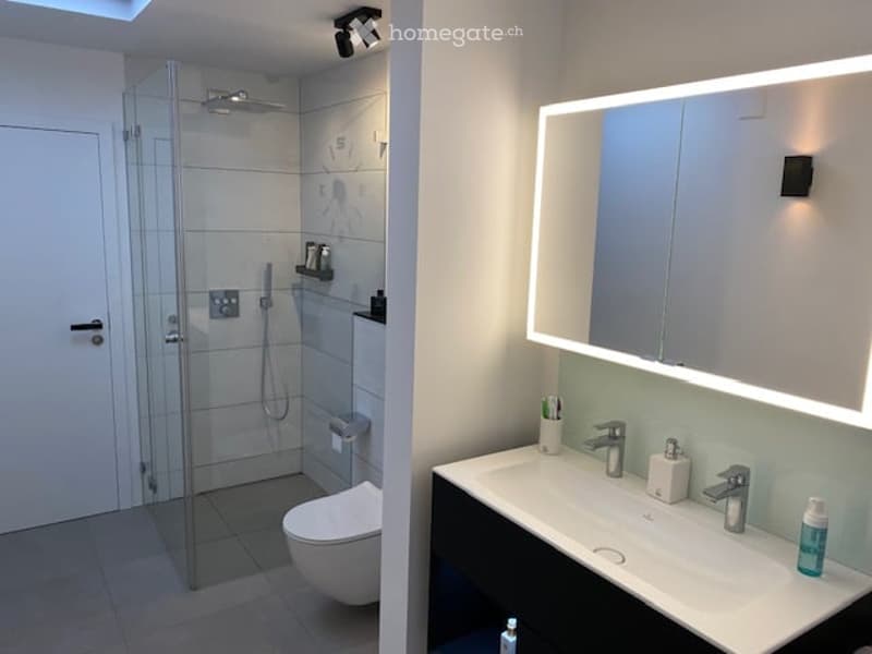 Modern bathroom with a glass-enclosed shower, toilet, and a double sink with a large illuminated mirror above it. The space features sleek, contemporary design elements and is well-lit.