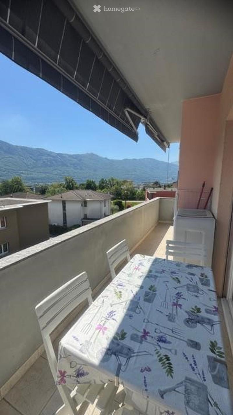 Balcony with outdoor furniture, scenic mountain view, clear blue sky, and surrounding houses.
