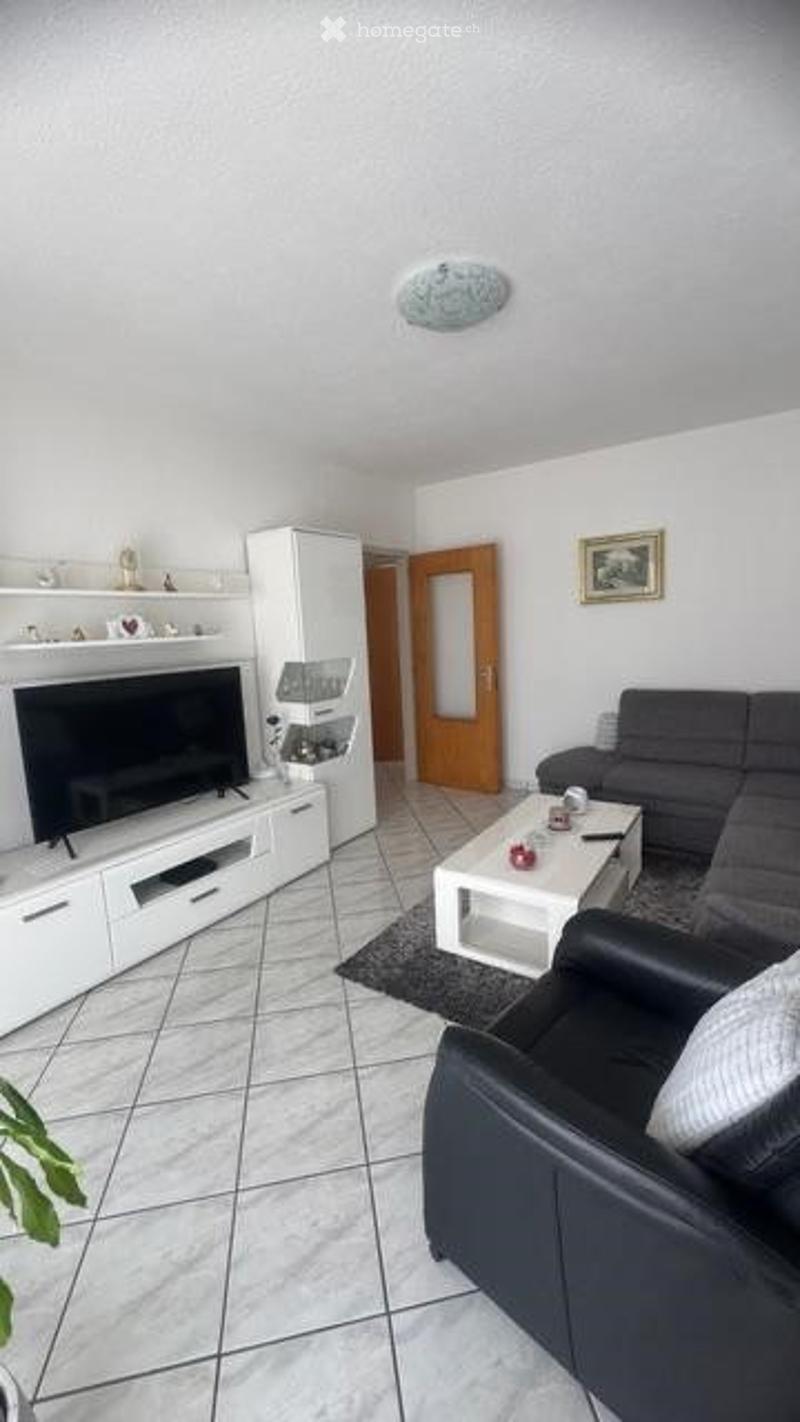 Modern living room with white tiled flooring, grey sectional sofa, white coffee table, flat-screen TV on white cabinet, decorative shelf, and a small plant in the corner.