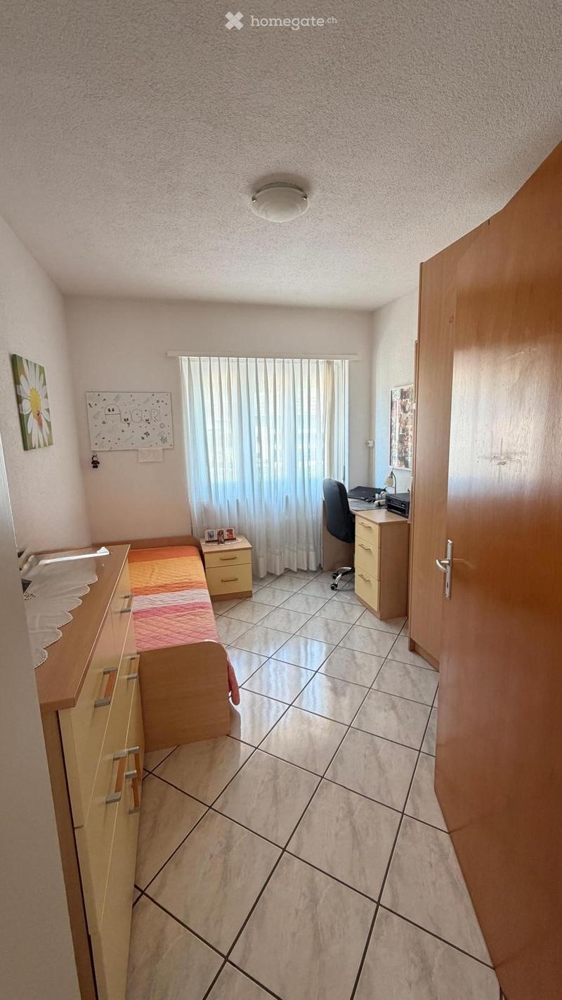 Cozy bedroom with tiled floor, bed with colorful bedding, nightstand, desk with chair, and cabinet.