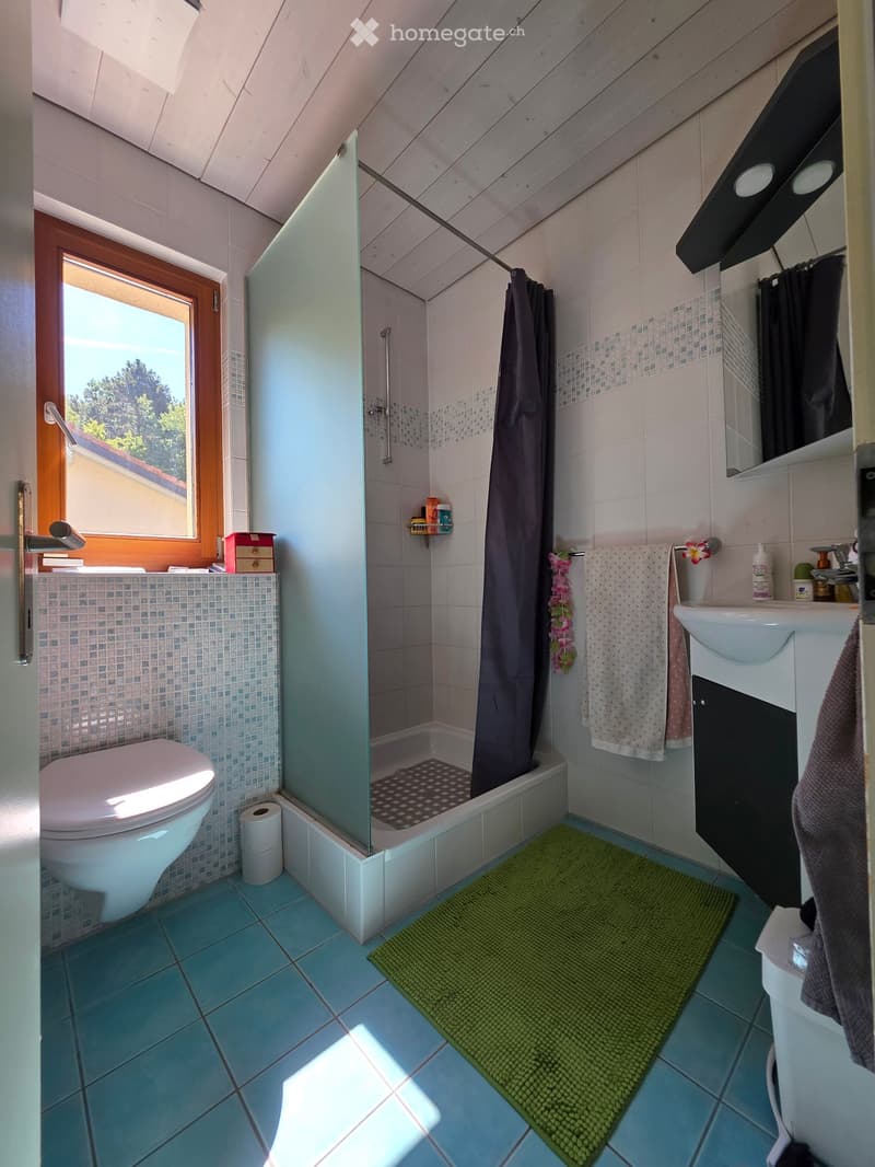 Bright bathroom with blue tiles, a toilet, a sink, a shower with a glass door, and a green bath mat. The room features a window with wooden framing, a towel rack with towels, and a small shelf with toiletries.