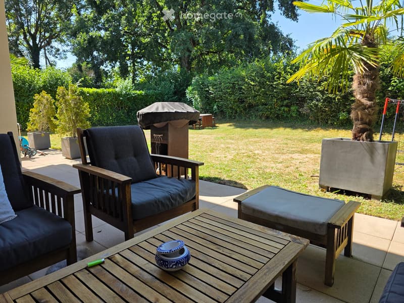 A well-maintained garden area with a wooden coffee table, two wooden chairs with dark cushions, a small ottoman, and a covered barbecue in the background. The area is surrounded by lush greenery and potted plants.