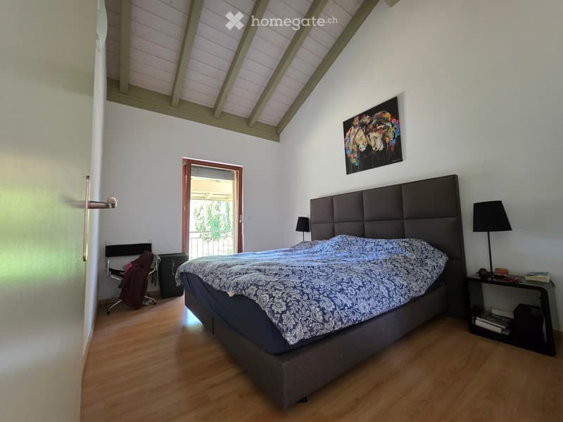 Cozy bedroom with wooden flooring, large bed with headboard, nightstand with lamp, chair, and a view to a balcony.