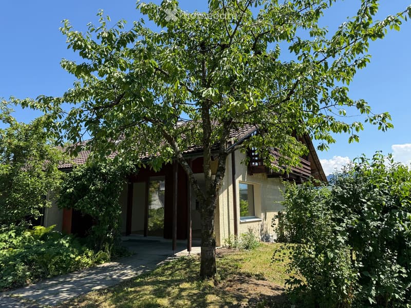 A two-story house with a tiled roof, surrounded by lush greenery and a large tree. The house features a balcony on the upper level and a pathway leading to the entrance.