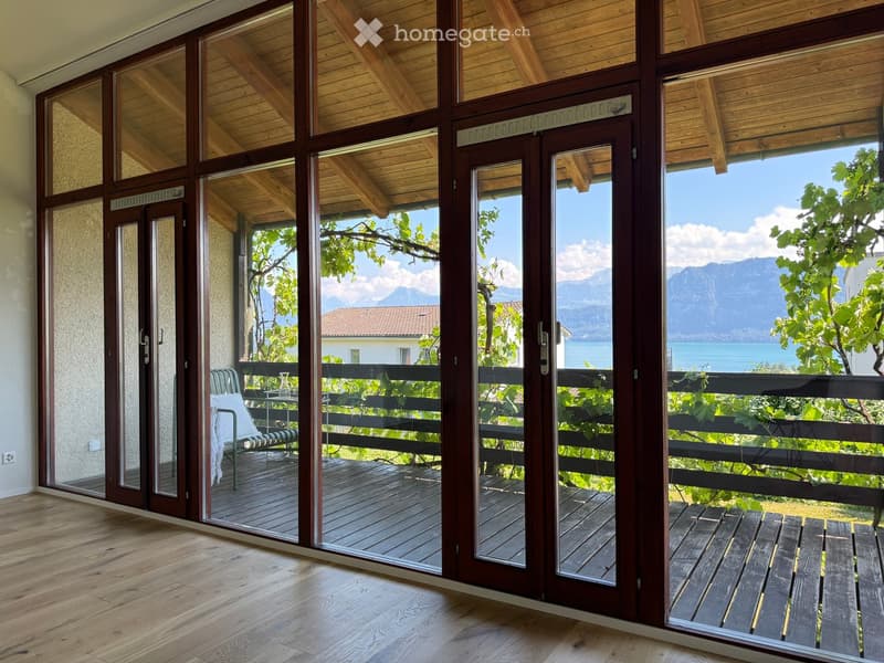 Open sliding glass doors lead to a wooden deck with a bench and a view of a lake and mountains.