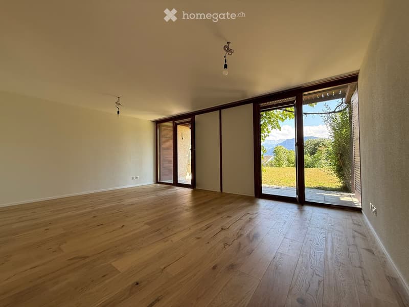 Empty room with wooden flooring, large sliding glass doors leading to a garden, and natural light from the windows.