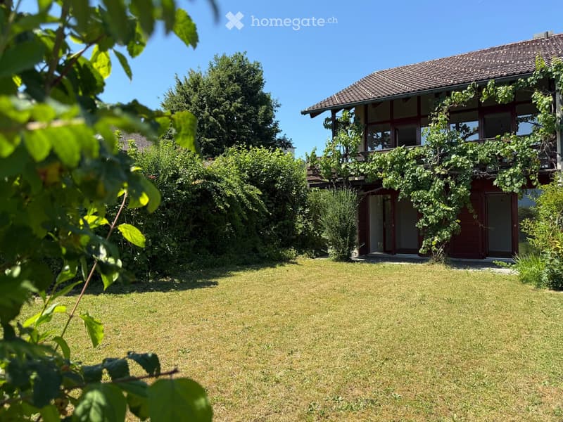 Lush garden with a well-maintained lawn, surrounded by trees and a two-story house with a tiled roof and covered balcony.