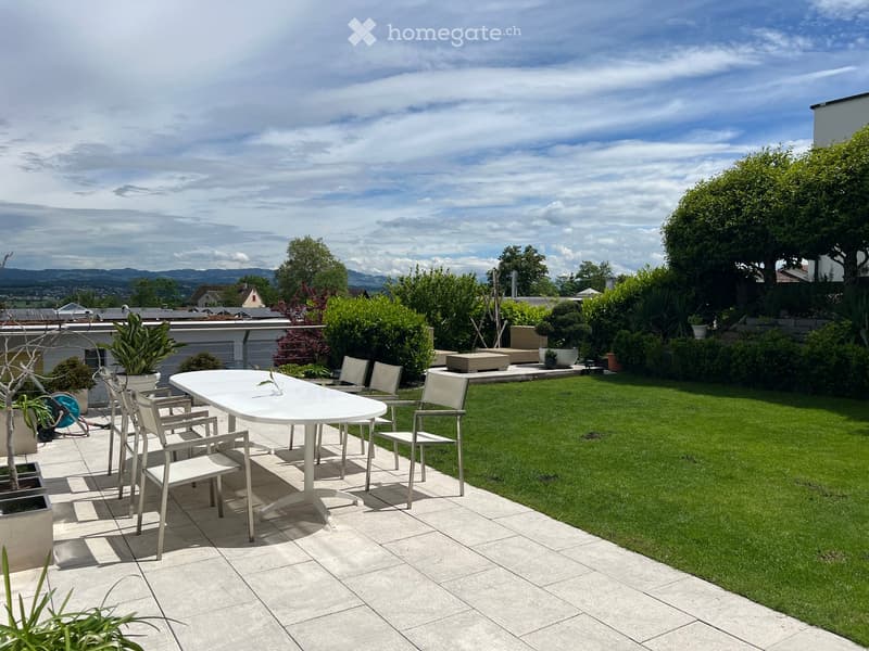 Well-maintained garden with a tiled patio area, white table and chairs, lush green lawn, and various potted plants.