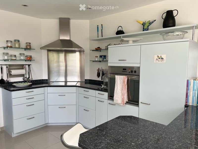 Modern kitchen with light grey cabinets, stainless steel appliances, granite countertops, and open shelving displaying jars, a teapot, and decorative items.