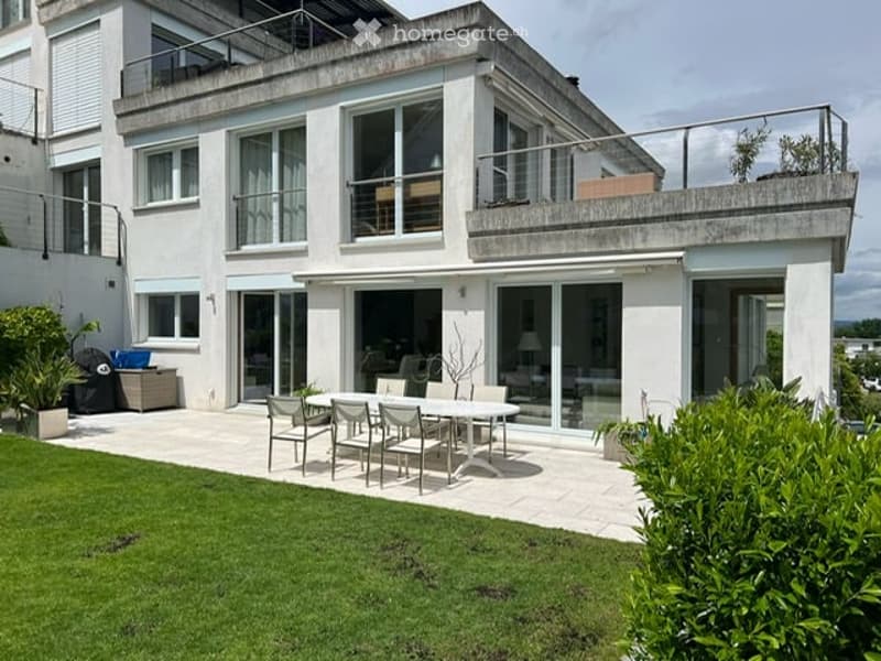 Modern house with a spacious outdoor patio area featuring a dining set, surrounded by greenery and featuring a balcony on the upper level.