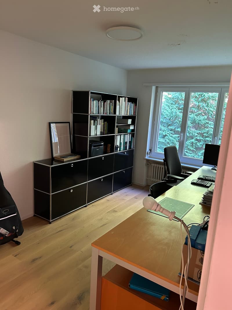 A well-lit office space with a wooden floor, large window, desk, chair, computer, keyboard, mouse, lamp, bookshelf, mirror, laptop, and printer.