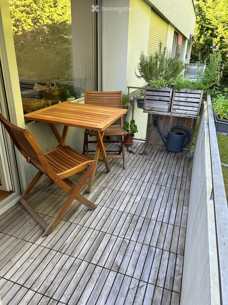 Balcony with wooden decking, a small wooden table, two wooden chairs, and a watering can.