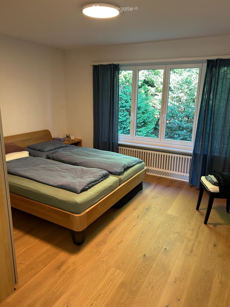 Cozy bedroom with wooden floor, large window with dark curtains, double bed with grey bedding, nightstand, small table, and chair.