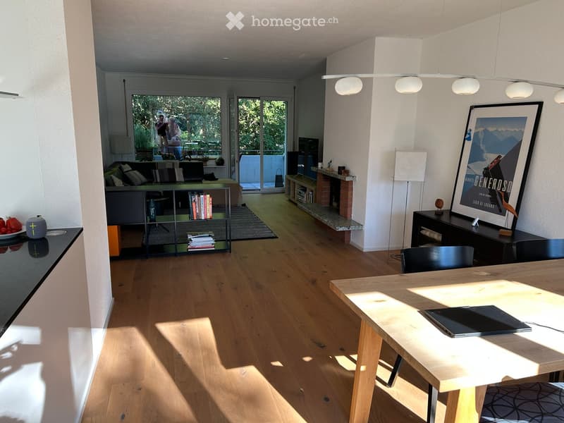 Bright living and dining area with wooden flooring, large windows, a sofa, a dining table, a bookshelf, and a fireplace.