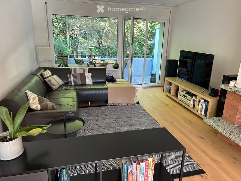 Modern living room with a green leather sofa, coffee table, bookshelf, TV, and fireplace. Large windows and sliding doors lead to a balcony.