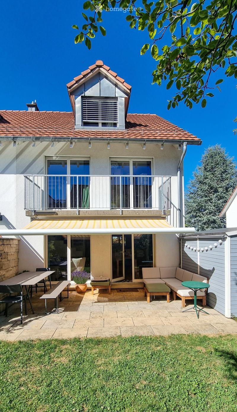 Outdoor patio area with a sofa, table, chairs, benches, and a flower pot, connected to a house with a balcony and covered awning.