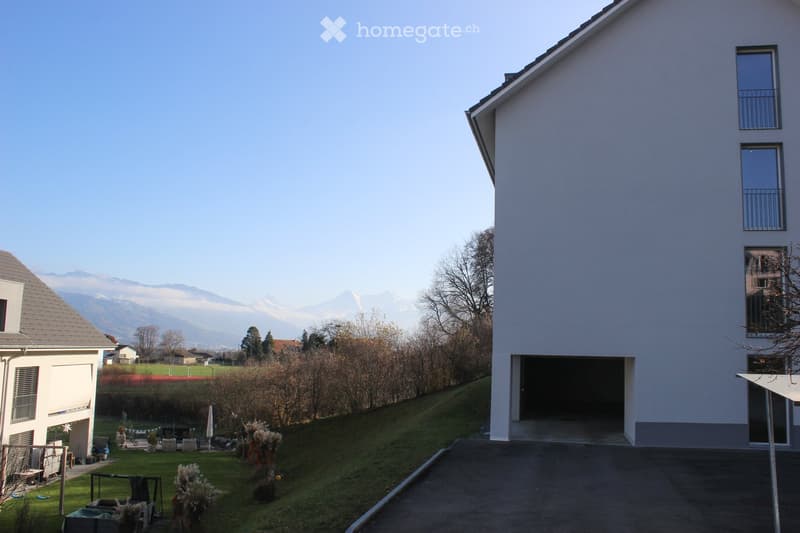 Modern two-story house with white exterior, large windows, and a garage entrance. The surrounding area includes a lawn, trees, and distant mountain views.