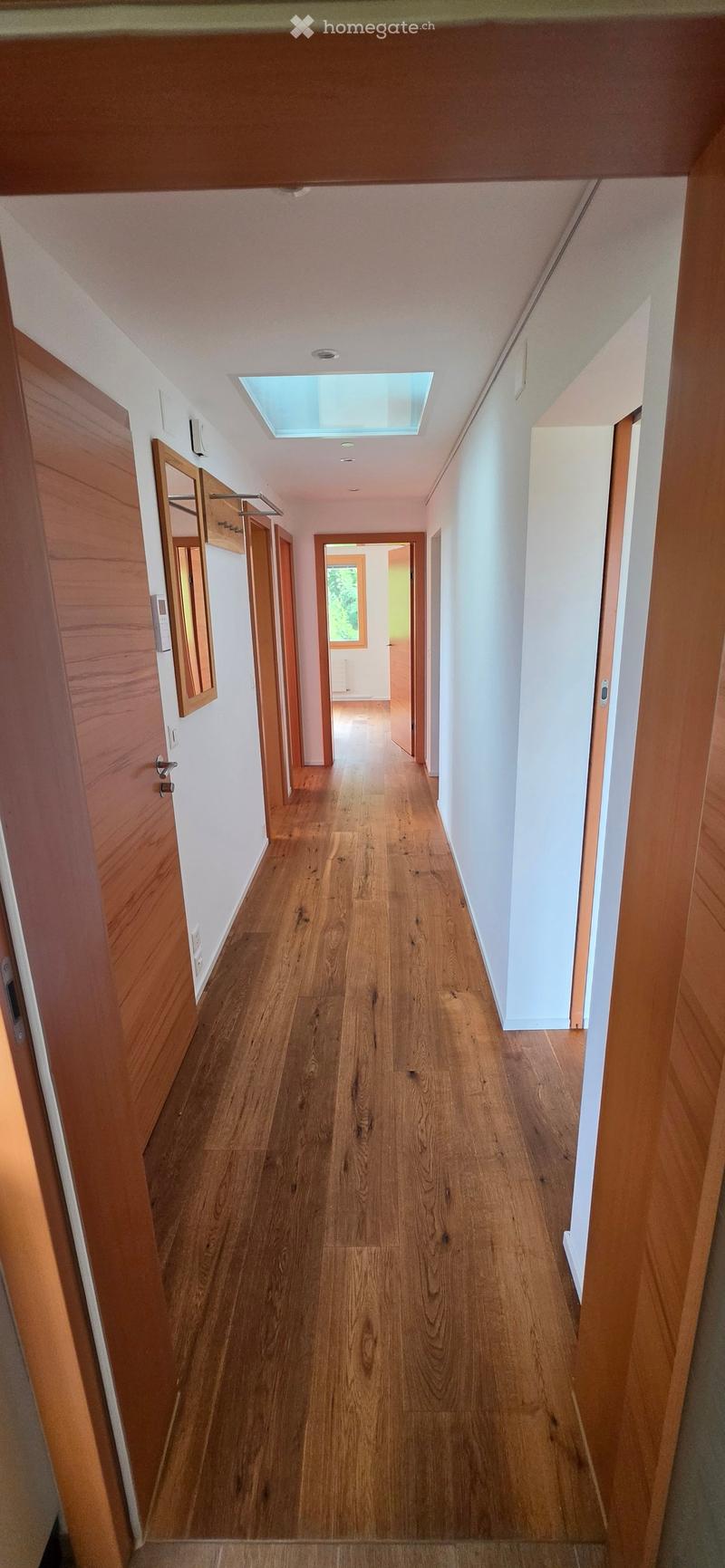 A long, narrow hallway with wooden flooring, white walls, and several doors on both sides. A skylight is visible at the far end, allowing natural light to enter.