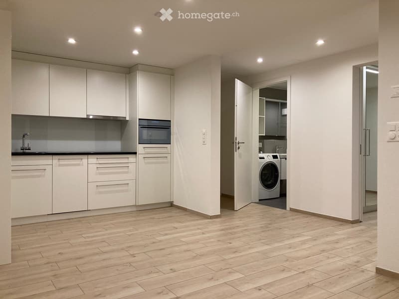 Modern kitchen with white cabinets, wooden flooring, and an open doorway leading to a laundry area with a washing machine.