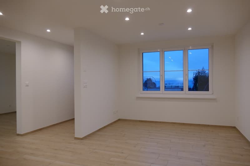 Empty room with wooden flooring, white walls, and large windows showing a view of the sea and trees.