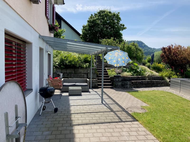 Outdoor patio area with a covered seating area, grill, umbrella, and steps leading to a garden.