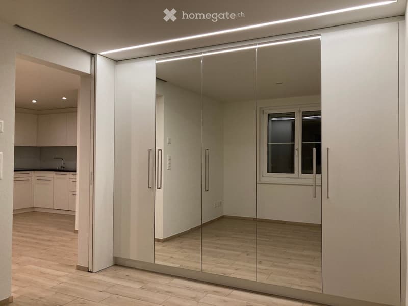Modern, minimalist room with light-colored wooden flooring, white walls, and a large mirrored cabinet with sleek handles. A small kitchen area is visible through an open doorway, featuring white cabinets, a sink, and a faucet.