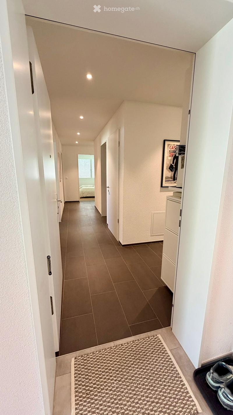 A modern hallway with tiled flooring, white walls, and recessed lighting. A small rug is placed near the entrance, and a pair of shoes is visible on a shoe rack. A framed poster is mounted on the wall, and a glimpse of a bedroom is visible at the end of t