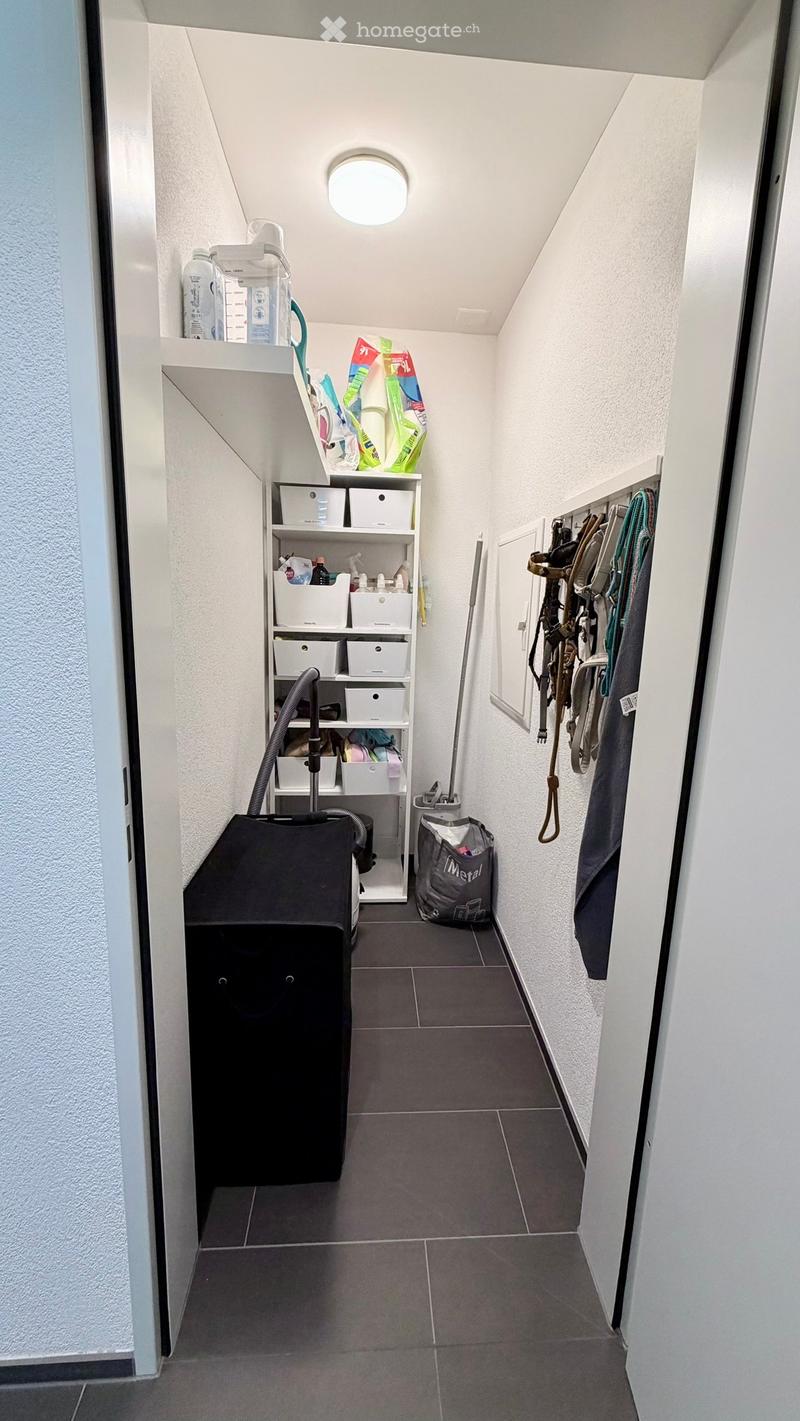 A narrow hallway with tiled flooring, white walls, and built-in shelving units holding cleaning supplies and storage boxes. A vacuum cleaner and a bag are placed on the floor, and a coat rack with various items is visible on the right side.