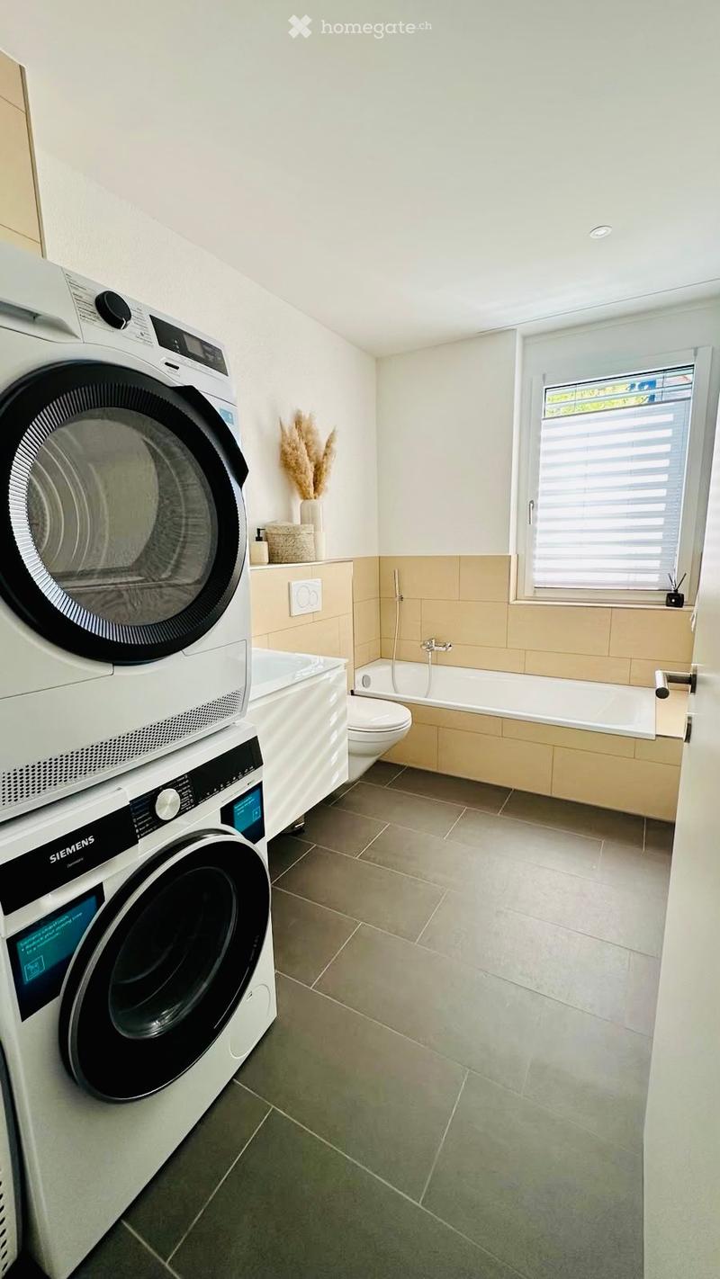 Modern bathroom with a washing machine and dryer unit, bathtub, toilet, and decorative vase on a shelf.