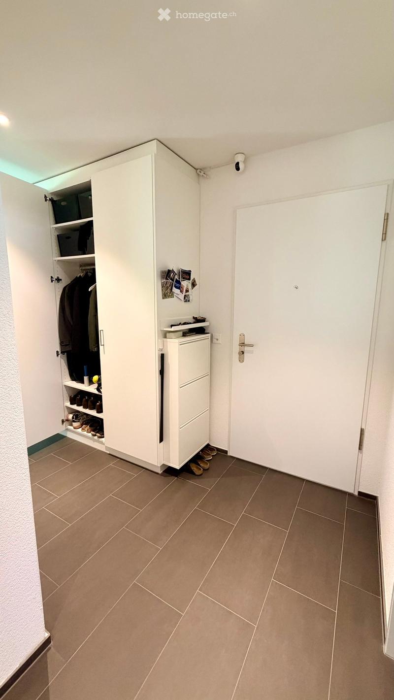 Empty hallway with tiled floor, white walls, built-in closet with shoes and clothing, small white cabinet, and a closed door.