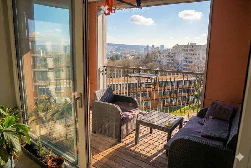 Balcony with wooden decking, wicker furniture including a sofa, chair, and small table, and potted plants. Offers a scenic city view.