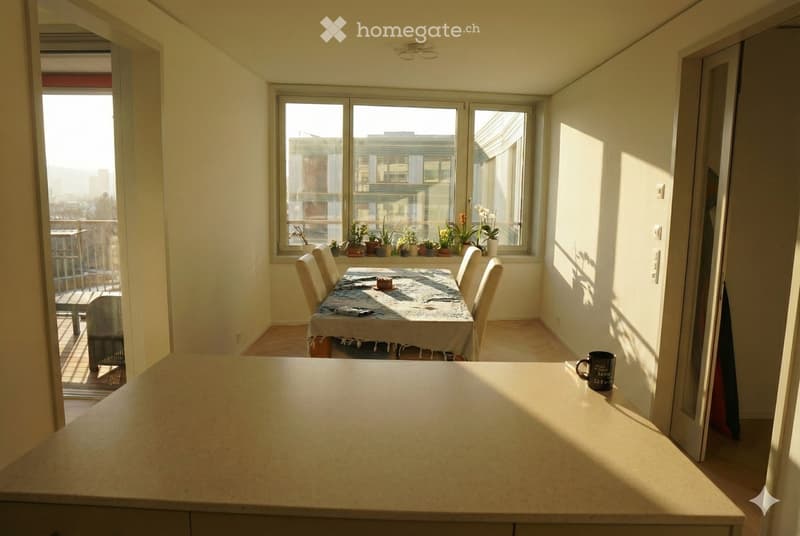 Bright dining area with a table, chairs, and potted plants on the windowsill. A coffee mug sits on a counter, and a balcony is visible through the open door.