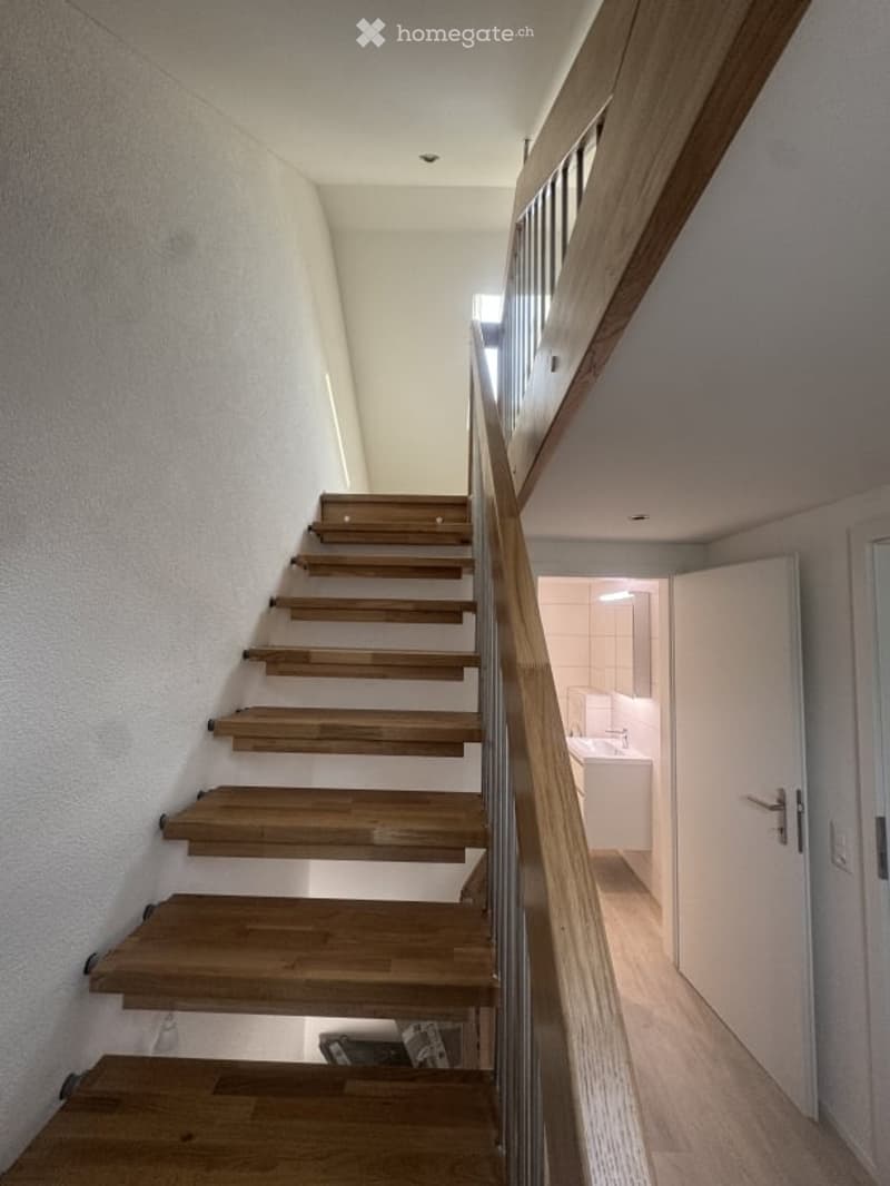 Staircase with wooden steps and a glass railing leading to a bathroom area with white tiles and a sink.